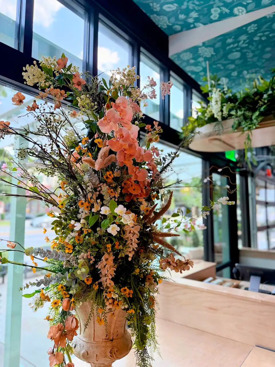 Towering faux arrangement of peach orchids, marigold violas and trailing greenery in a stone urn by a sunlit restaurant window